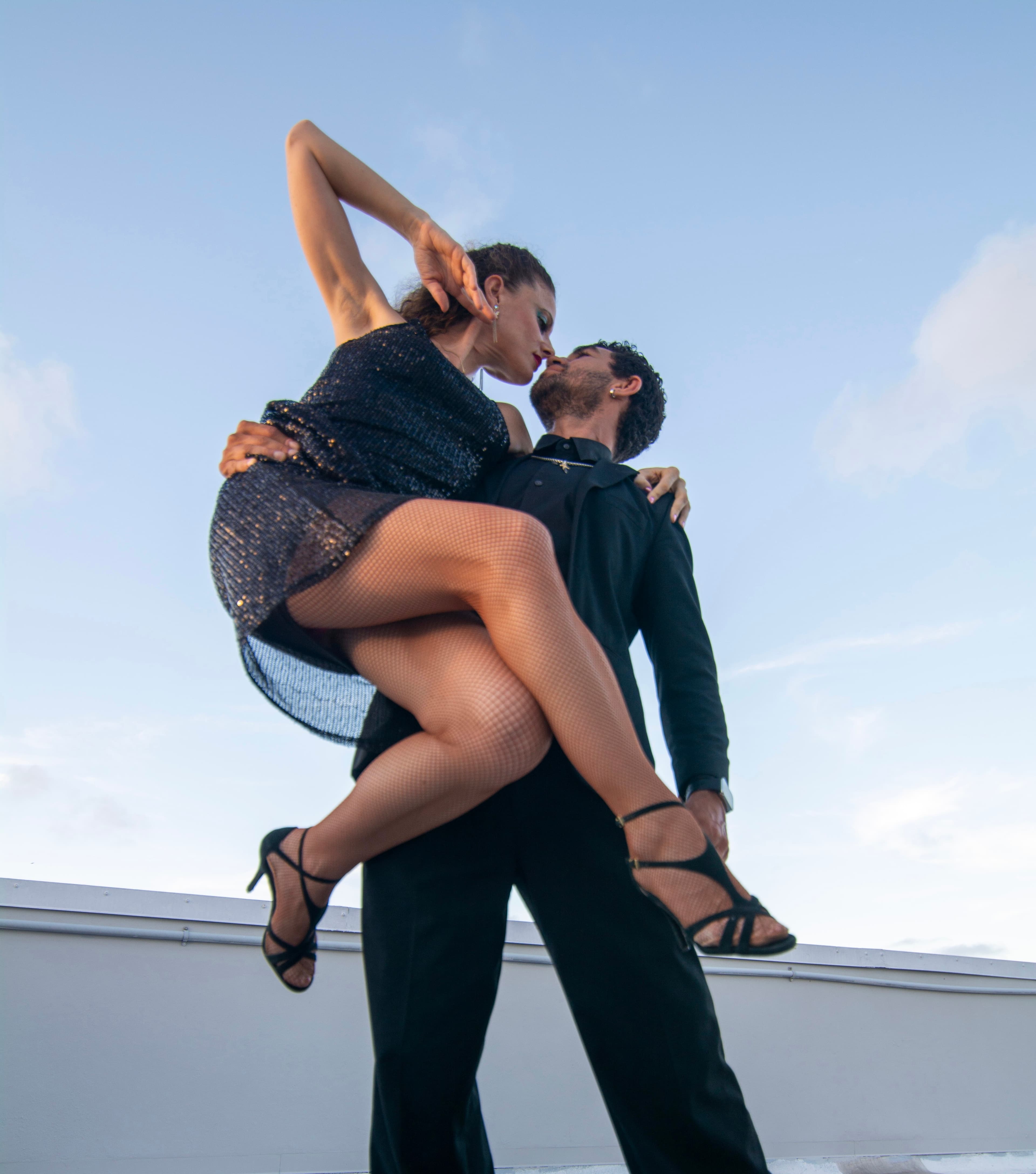 Man in black suit lifts woman in sparkly dress during a dramatic rooftop dance.