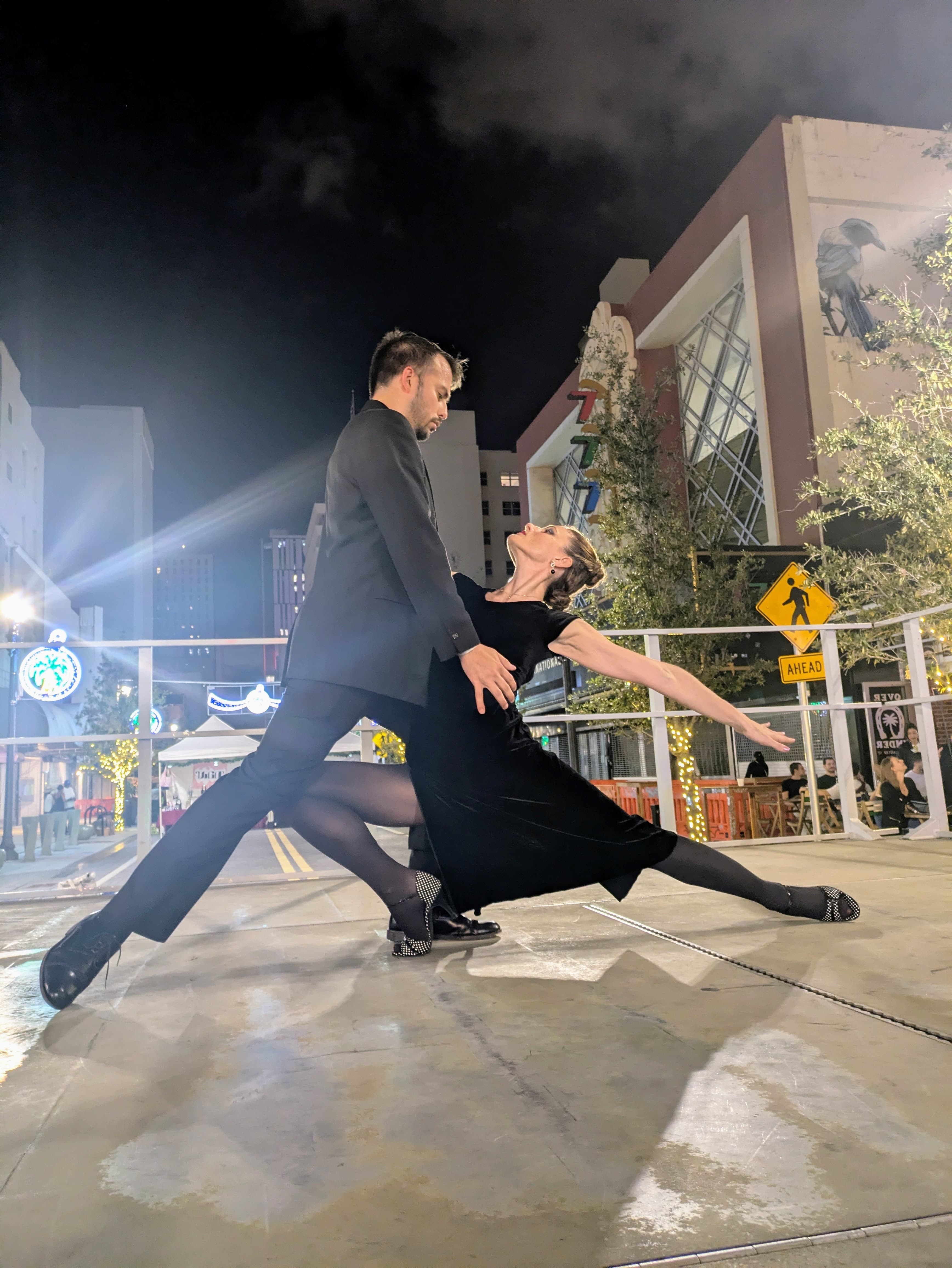 Couple in black formal attire performs a dramatic dance pose on a night city street.