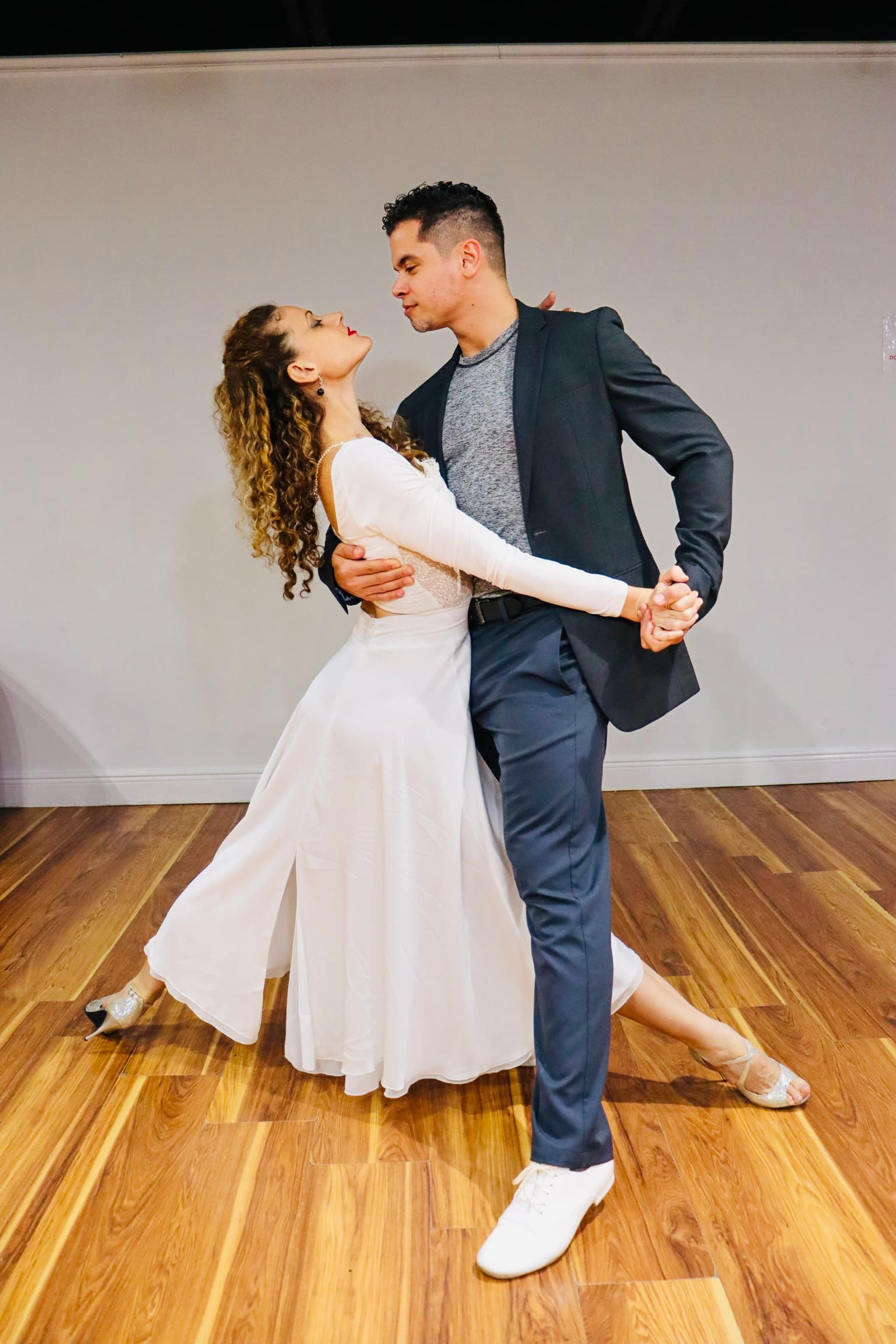 Man in a blazer and woman in a white dress performing an elegant dance pose.