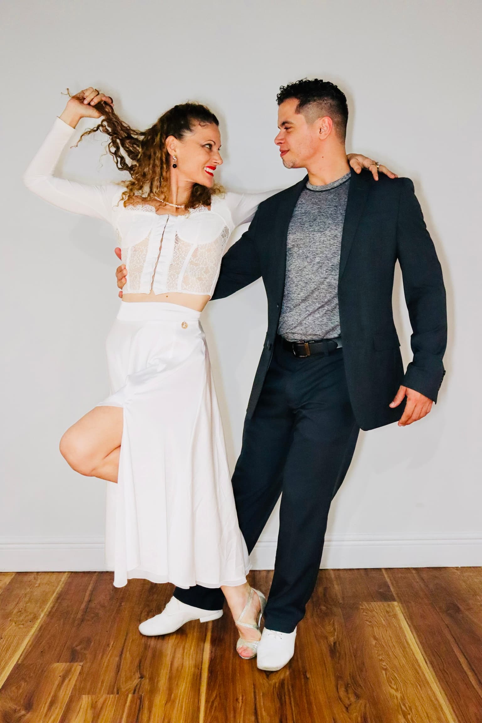 Man in dark blazer and woman in white lace outfit posing playfully against white wall.