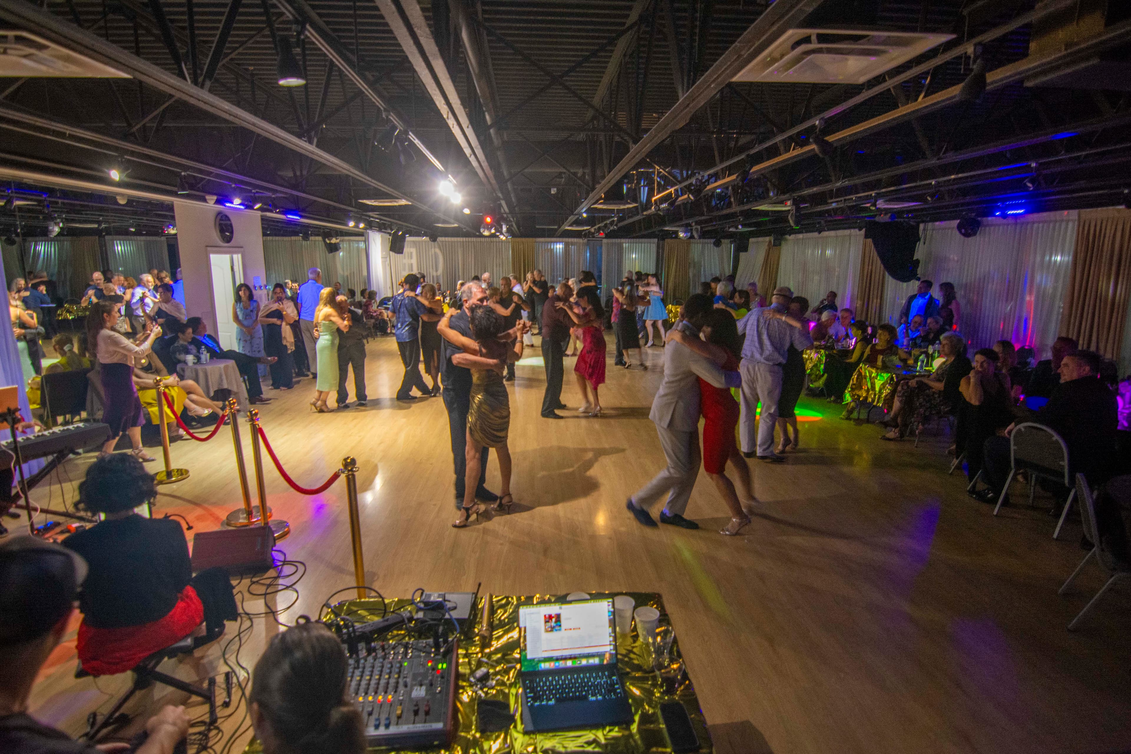 Couples ballroom dance on a wooden floor in a dimly lit studio with DJ equipment foreground.