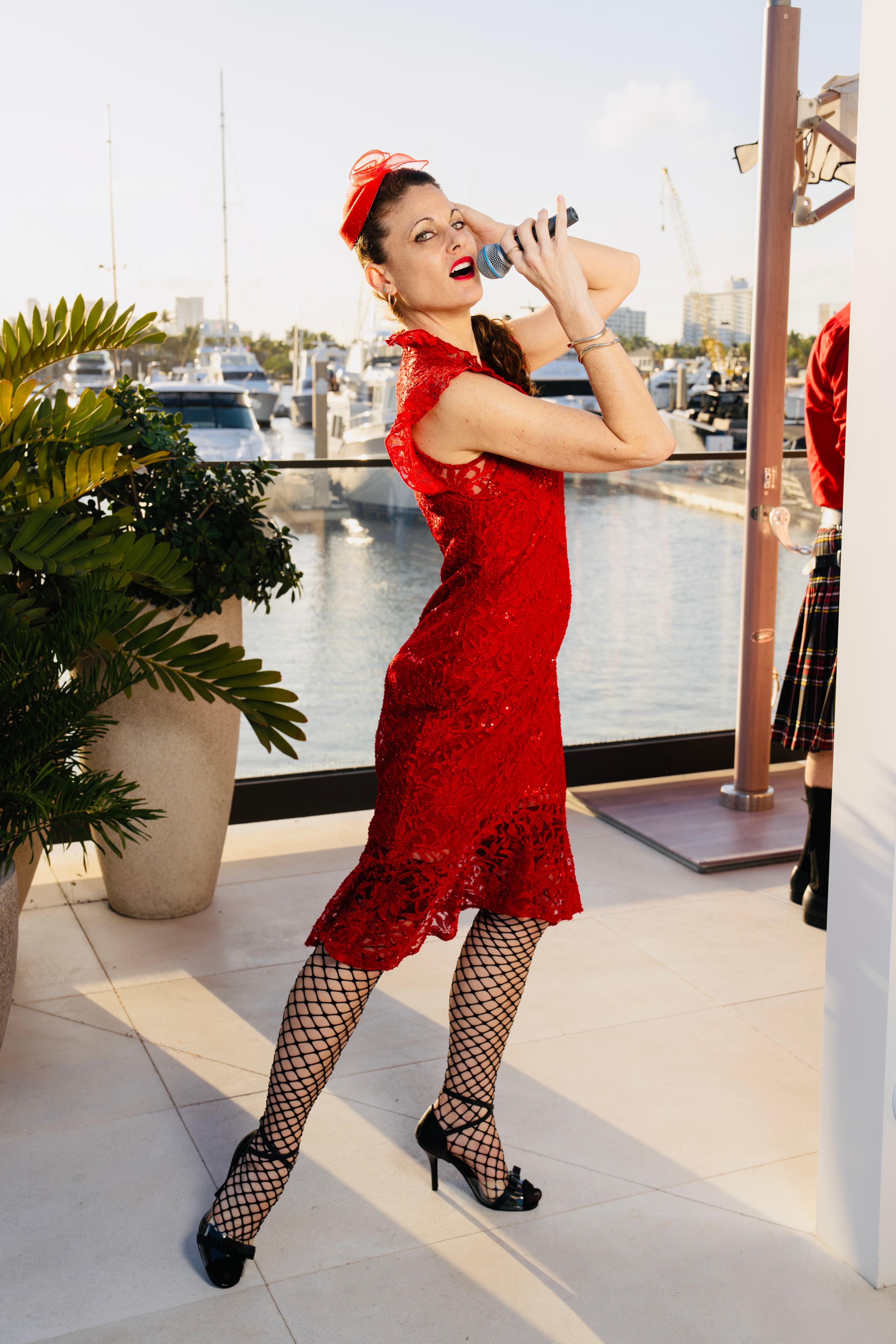 Woman in a red lace dress and fishnets singing into a microphone at a marina.