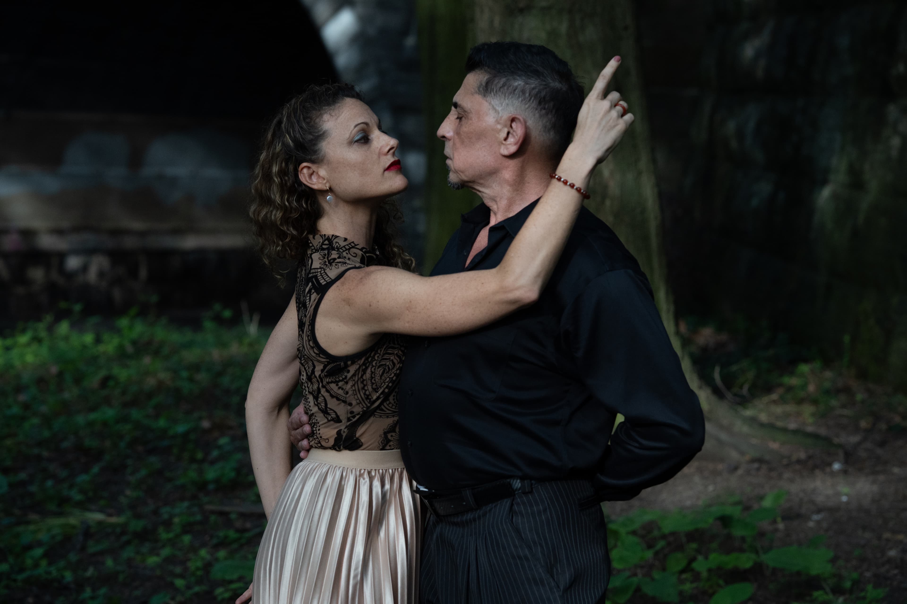 Couple in formal attire pose dramatically, gazing intensely at each other in a dark forest.