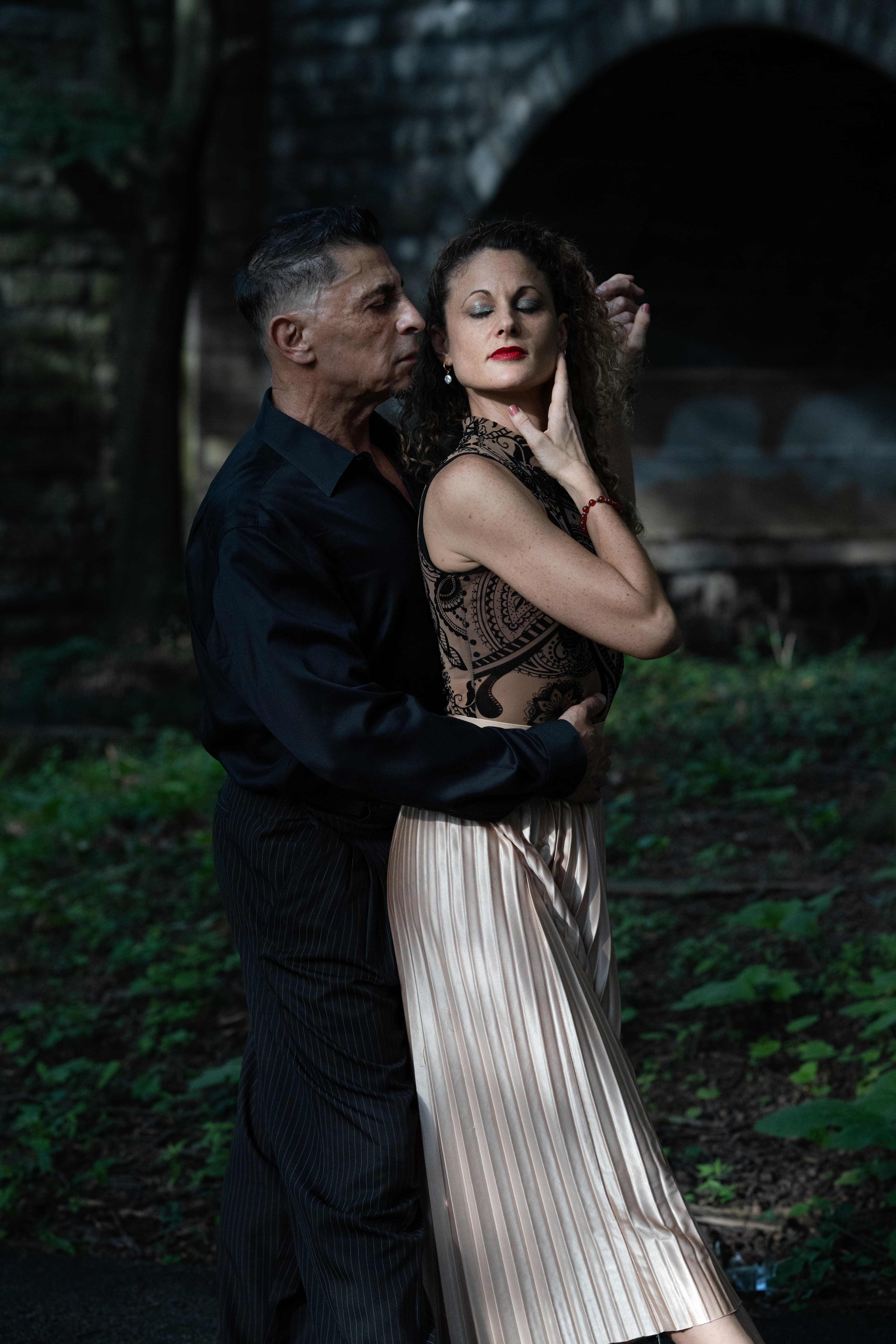 Man in black and woman in gold skirt perform an intimate tango pose outdoors.