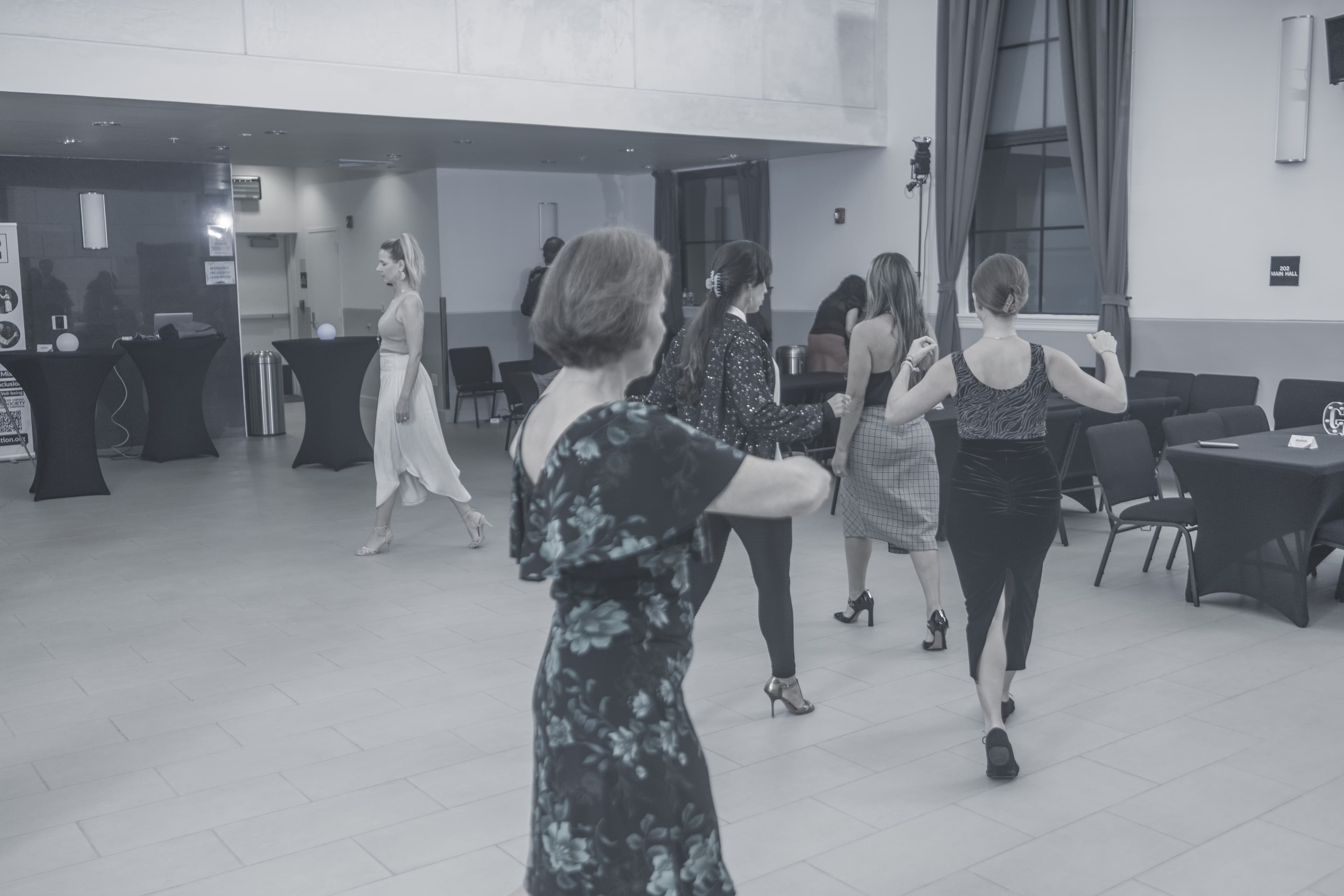 Several women in dresses dance and walk across a large, open indoor event space.
